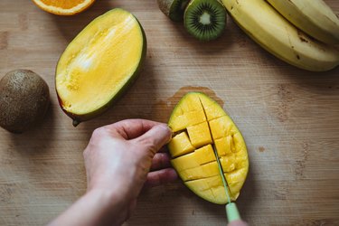 an overhead photo of a person's hand cutting a green unripe mango next to a kiwi and bananas