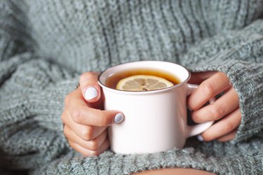 a close up of hands holding a white mug of hot ginger tea with lemon to debloat fast