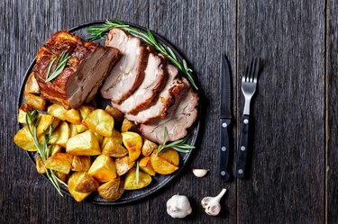 Plate of sliced, roasted boneless pork sirloin roast with potatoes and fresh rosemary on top on a black plate beside a knife and fork on a dark gray wooden table.