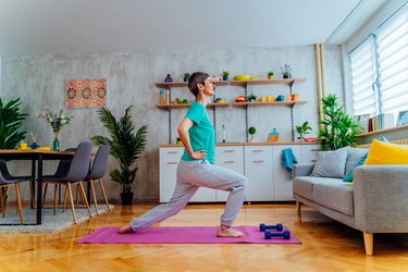 older woman doing the Pilates scooter lunge exercise on a pink yoga mat in her living room