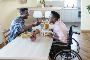 Person in wheelchair seated across from partner eating breakfast at home in the dining room