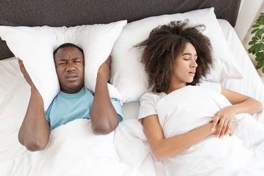 a man in bed with a pillow around his ears to drown out his wife's snoring, which is a sign of sleep apnea