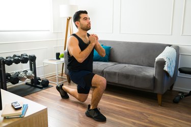 Adult athlete doing split squats in living room wearing black tank top and shorts.