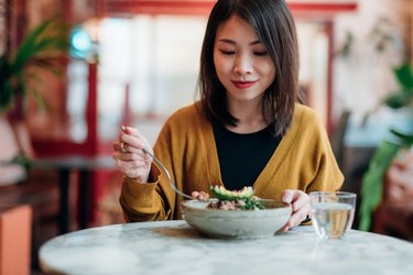 a person wearing a yellow sweater over a black shirt is eating a green salad in a gray bowl at restaurant with a marble table and a glass of water