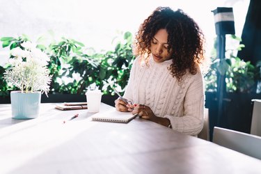 Young ethnic woman writing in notebook in cafe