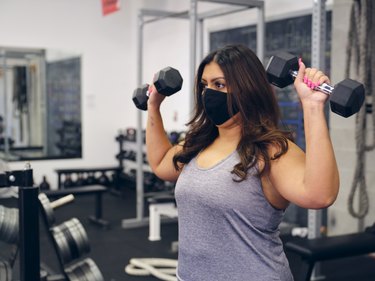 Plus-size woman wearing a mask and strength training with dumbbells at the gym