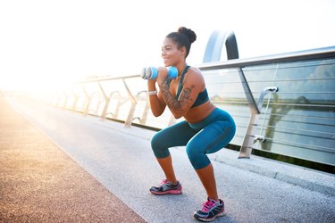 fit woman wearing blue workout leggings and working out with a pair of blue dumbbells outside