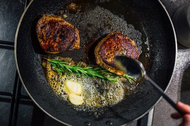 Frying pan with two steaks on the stove with butter and herbs to be cooked in the oven to finish