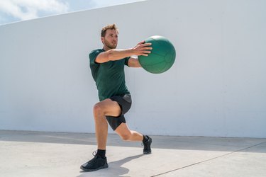 Person in workout attire doing lunges and torso rotations with a medicine ball outside.