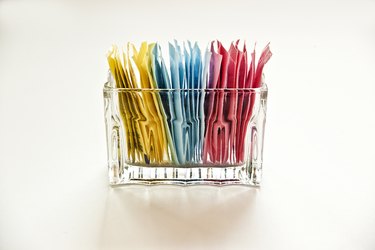 packets of yellow, blue and pink artificial sweeteners in a clear glass container on a white background