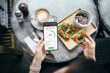 Overhead view of person's hands using mobile app on smartphone to track their daily diet meal plan and calorie intake while eating sourdough toast with smashed avocado and cherry tomatoes in a restaurant