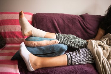 Injured hispanic woman elevating bandaged sprained foot on a pile of pillows on the sofa