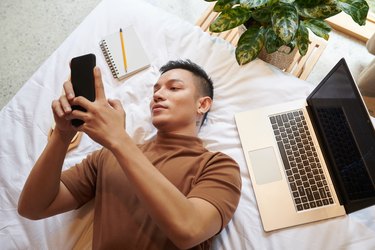 Person looking at phone while lying on bed next to computer