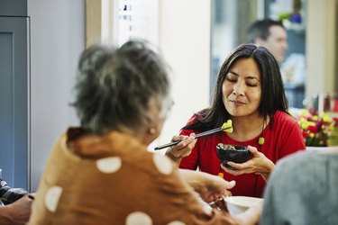 Person with long black hair and a red shirt eating food with chopsticks and looking pensive
