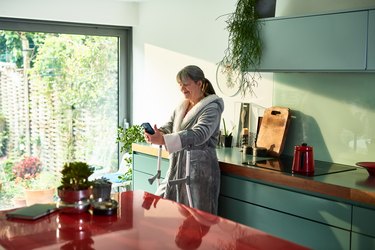 a person with a gray ponytail and forearm crutches wearing a gray bathrobe and standing in their kitchen smiling at their smartphone