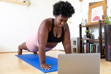 Person in a black sports bra and pink shorts doing a plank on a blue yoga mat at home, as an exercise to reduce sagging breasts