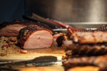 Canadian bacon, also known as peameal, for sale, grilled and cooked, being ready to be cut on a wooden plank, surrounded by other cooked meats.