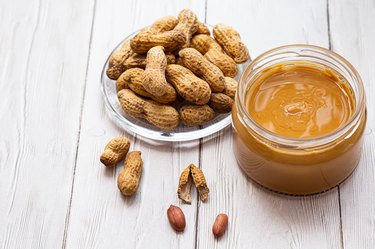 A jar of peanut butter with a bunch of peanuts on a white wooden table. Homemade peanut butter, natural, healthy food. The modern wellness and vegan concept.