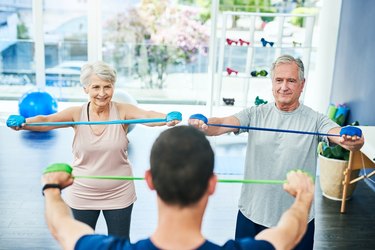 older couple doing band pull-aparts with their trainer at a gym