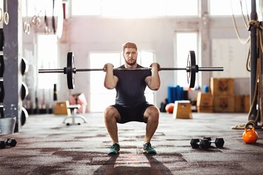 Man doing a barbell squat in a gym with dumbbells behind him