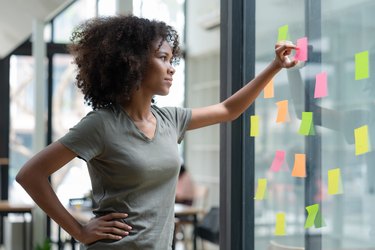 a young adult with brown curly hair wearing a gray t-shirt stands with their hand on their hip in an office looking at an arrangement of sticky notes taped on a window