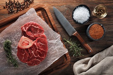 Raw calf shank beef with bone on white cooking paper and wooden cutting table. Dercorated with herbs, spices and chef's knife.