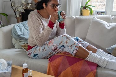 Person with a cold, sitting on white couch in the living room while drinking tea. Medicine and tissue paper on table.