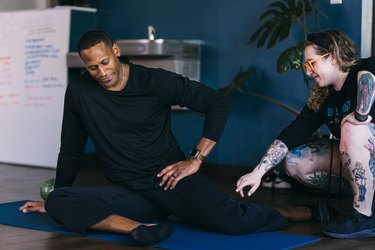 a fitness instructor with long hair and tattoos helps a middle-aged fit man do hip flexor stretches on a mat