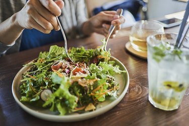 close view of a person's hands tossing a salad on a plate, next to a glass of seltzer water with green herbs