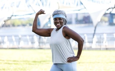 older adult wearing a blue headband, tank top and leggings flexes biceps outside