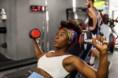 Person wearing white sports bra and blue leggings strength training in the gym using dumbbells