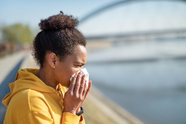 black woman wearing a yellow hoodie sneezing and blowing nose into a tissue while exercising outdoors