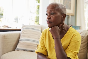 an older adult wearing a yellow shirt sits on a tan couch at home looking depressed