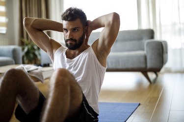 Person with short hair and beard doing a sit-up on an exercise mat at home in living room.