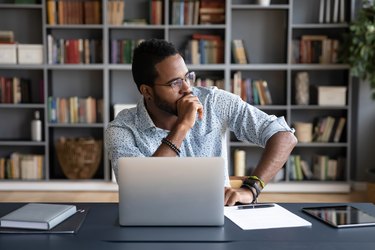 a person wearing a light blue button down shirt and glasses sits at a desk in front of a laptop with a bookcase in the background looking distracted from brain fog and looking off into the distance