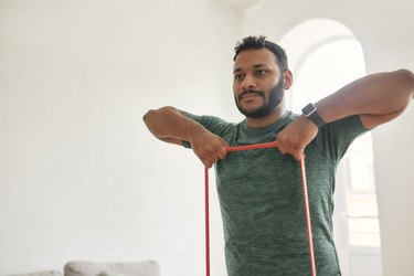 Work out indoors. Cropped shot of young active man looking away, exercising with resistance band during morning workout at home. Sport, healthy lifestyle