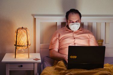 Adult man in a medical face mask N95 on the bed with a computer