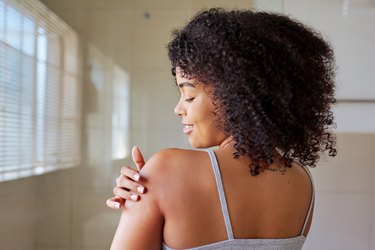 Photo of a person with curly hair smiling while applying lotion to their shoulder and wearing a light grey tank top
