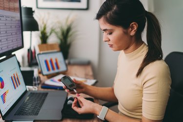 a person with long dark hair in a ponytail wearing a tan shirt tests blood sugar with a finger prick while sitting at a desk at home in front of a laptop showing graphs and charts