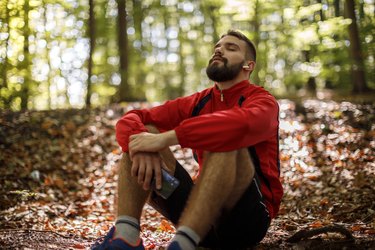 Portrait of a relaxed young man with bluetooth headphones sitting in a forest and holding his breath