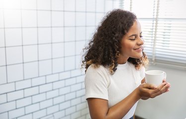 年轻美丽的非裔美国妇女手握咖啡杯的肖像早上冬天的时间在白色的卧室。快乐开朗放松。唤醒舒适hygge常规大学生活方式的概念。