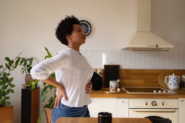 Person in a white long sleeve shirt and jeans with a short hair cut standing in their kitchen with one hand on their lower back and the other hand on their stomach wincing in discomfort.