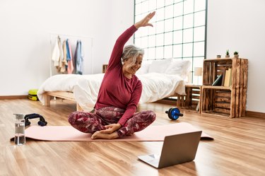 Middle age grey-haired woman having stretching online class sitting on the floor at bedroom.