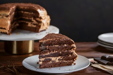 A slice of homemade brownie chocolate cake on a white platter. The layer cake is covered with butter icing with condensed milk and chocolate chips. White plates and forks decorate the table.