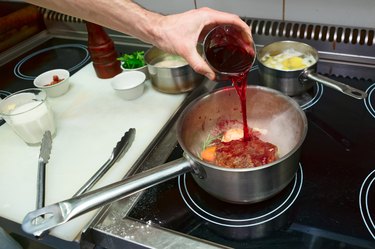 Hand pouring red wine into a pot of cooking meat and vegetables
