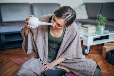 young woman sitting in a living room and using neti pot, as a natural remedy for sinus infection