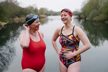 Two older adult swimmers in one pieces and swim caps laughing in front of a lake