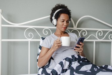 a woman drinking coffee and listening to a mental health podcast in bed