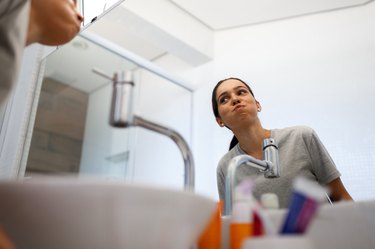 a person wearing a grey t-shirt with brown hair in a ponytail swishing mouthwash at the bathroom sink