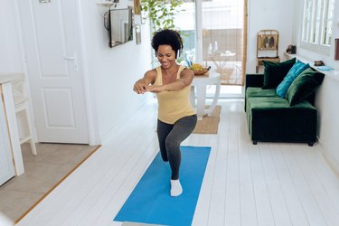 African American woman doing lunges on yoga mat in living room while wearing headphones